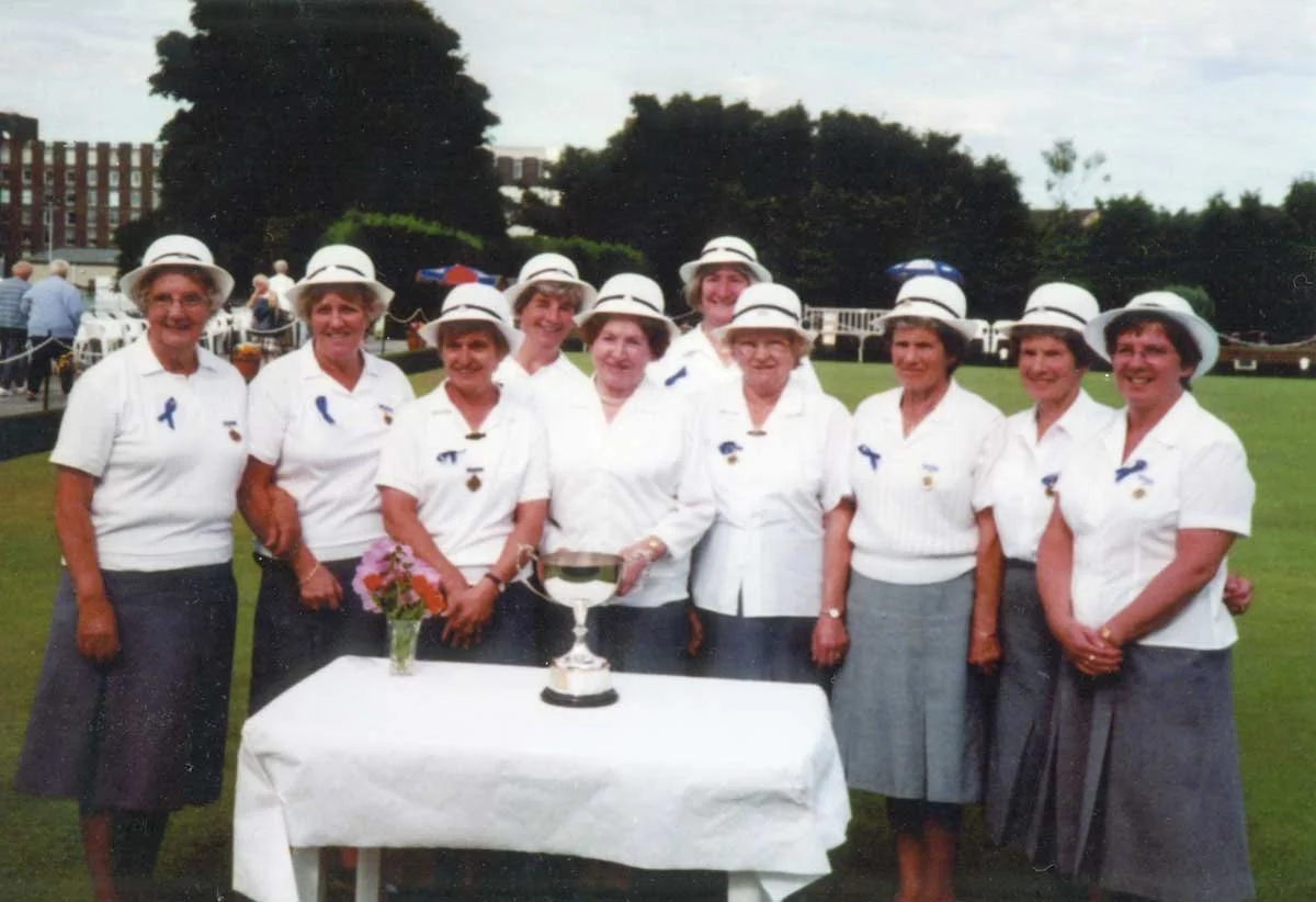 Group of women dressed in white shirts and gray skirts, wearing white hats, standing outdoors on a grassy area, smiling, with a table in front with a trophy and a small vase of flowers, possibly at a golf or social event.