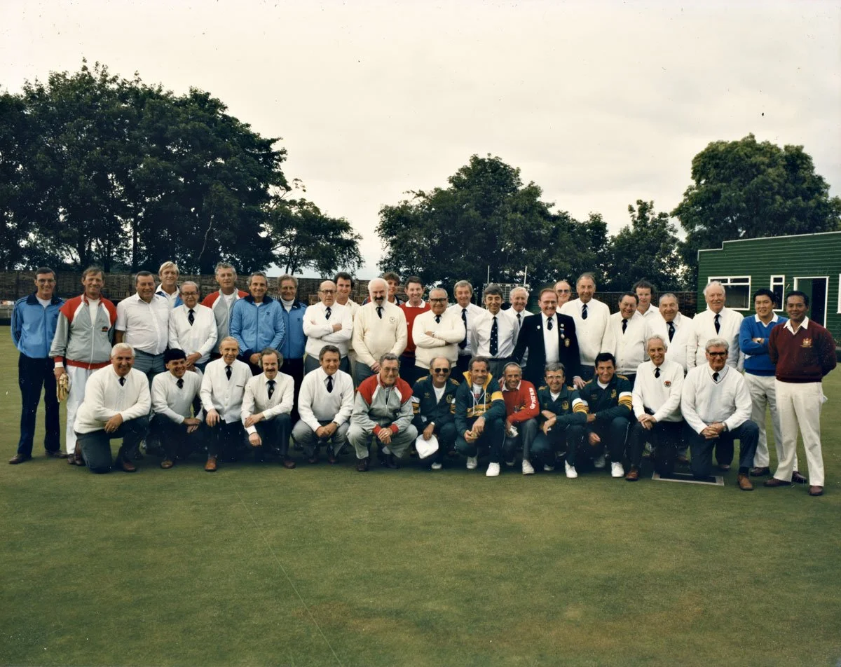 Group of men on a cricket field, dressed in cricket and formal attire, posing for a photo.