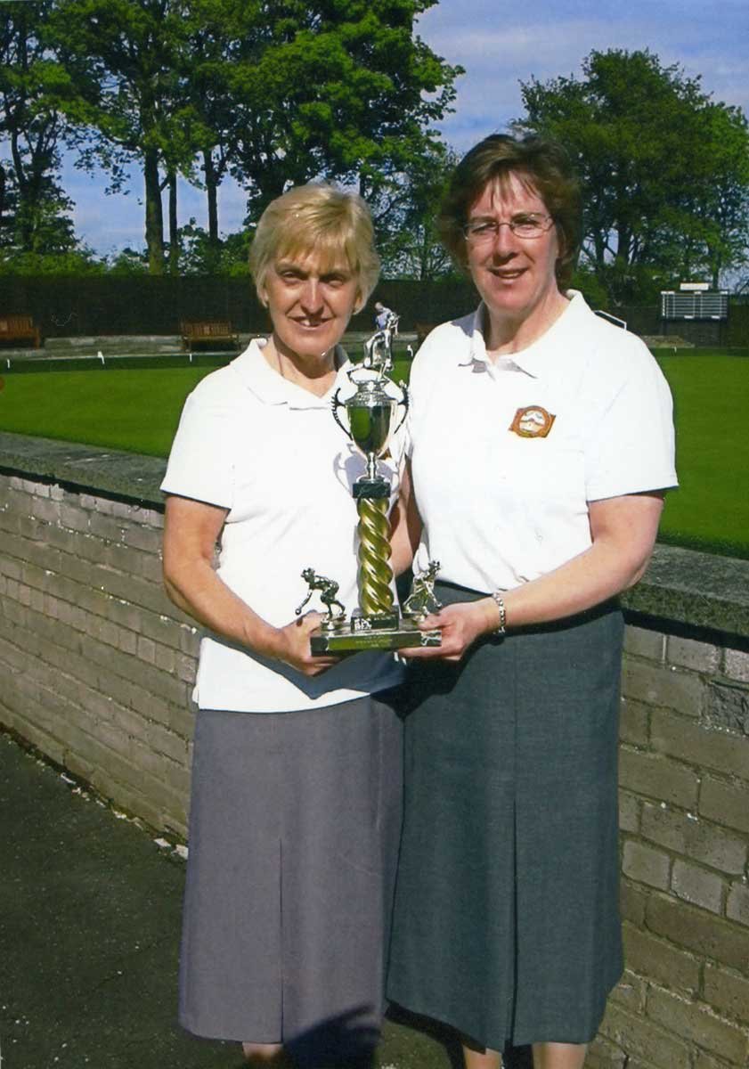 Two women in white shirts and gray skirts holding a trophy together outdoors on a sunny day, with trees and a grassy area in the background.