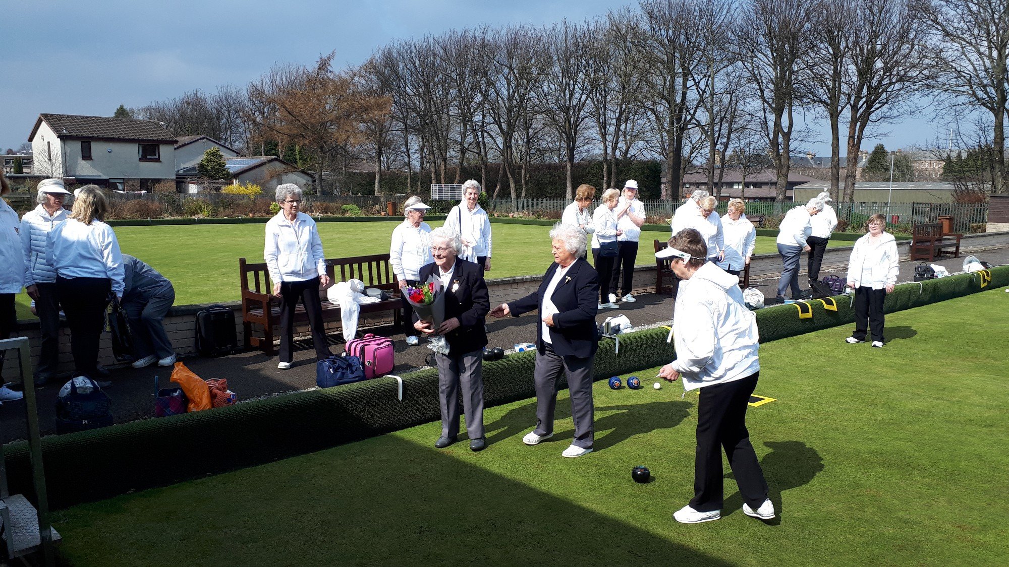 Group of elderly women playing lawn bowls outdoors on a sunny day, some preparing to bowl, others watching, with trees, houses, and blue sky in the background.