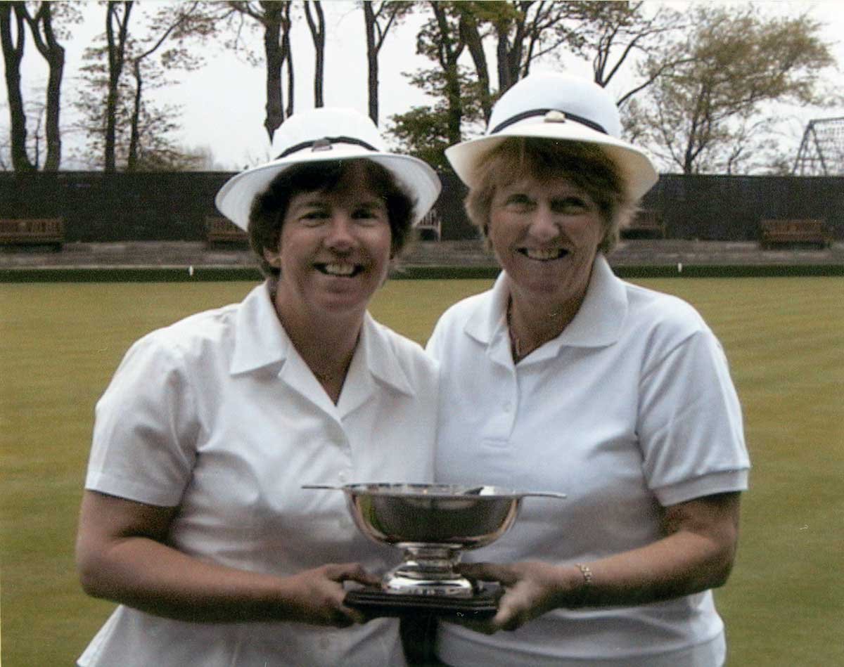 Two women in white golf shirts and white hats holding a silver trophy on a golf course, smiling at the camera.
