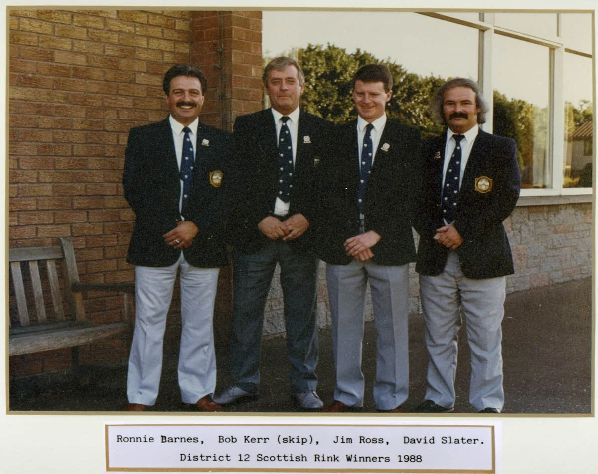 Four men standing outdoors against a brick wall and a window, dressed in matching blazers and ties, posing for a photo. The caption indicates they are Ronnie Barnes, Bob Kerr (skipping), Jim Ross, and David Slater, winners of District 12 Scottish Rin