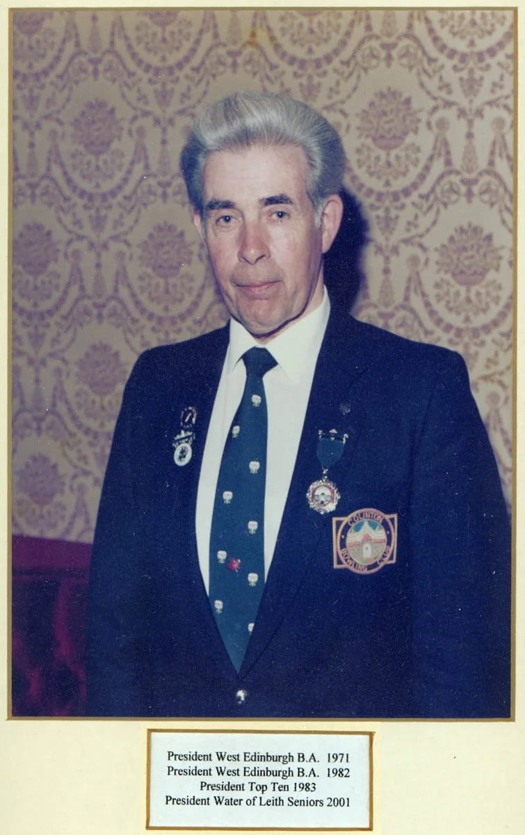 A man with silver hair wearing a black blazer, white shirt, and a black tie with a pattern of small white skulls. He has several pins and medals on his blazer. The background features patterned wallpaper. The image includes a label with his titles an