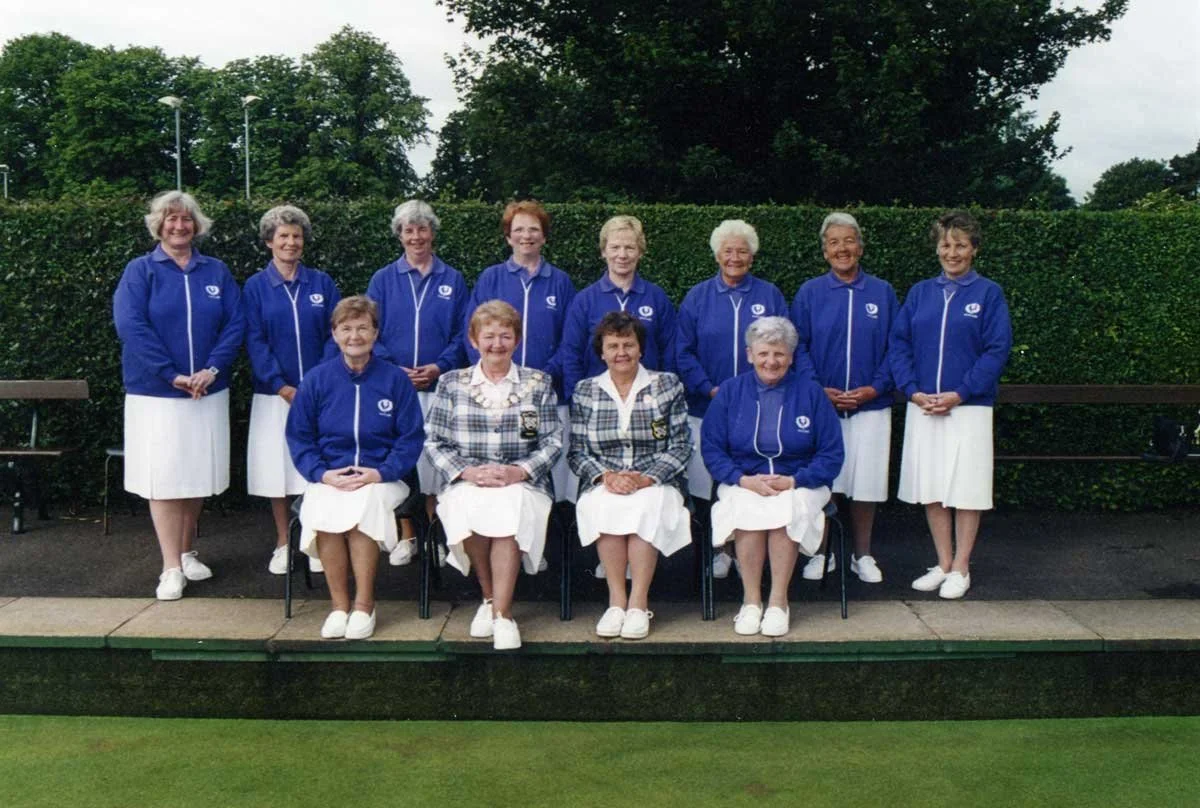 A group of eleven women, some sitting and some standing, wearing matching blue jackets with a white emblem and white skirts, posing outdoors on a lawn with bushes and trees in the background.
