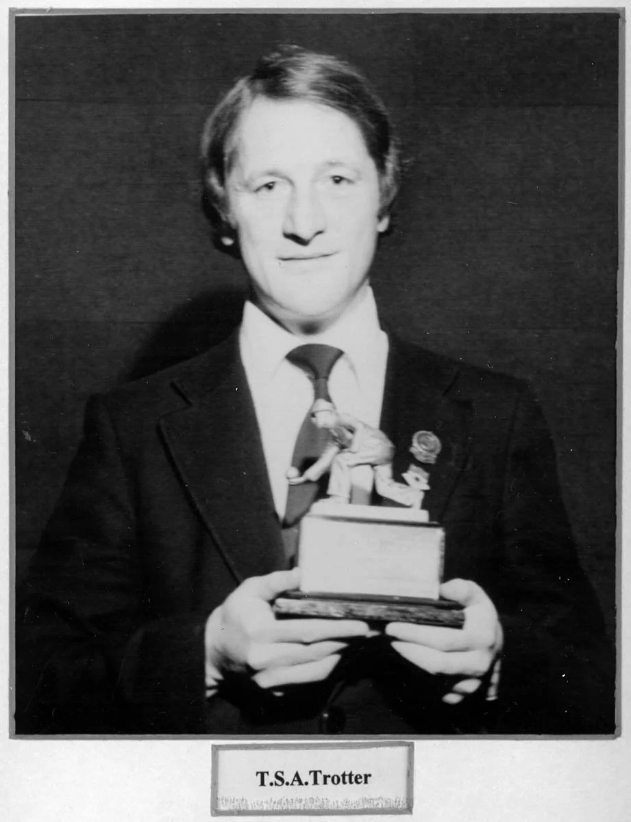 Young man in formal suit holding a trophy or award with a figurine of a person in a tennis stance, labeled "T.S.A. Trotter."