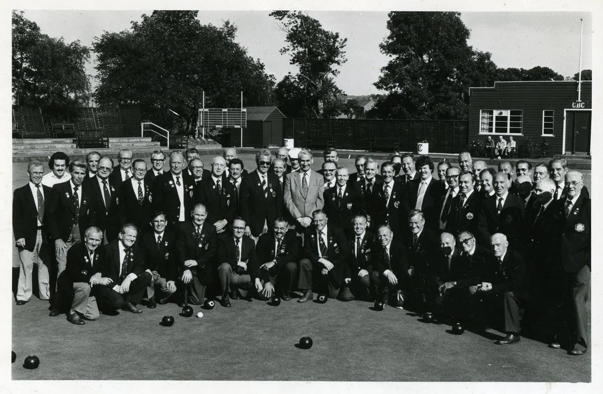 Group of men dressed in suits on a lawn playing lawn bowls, with trees, fencing, and a small building in the background.