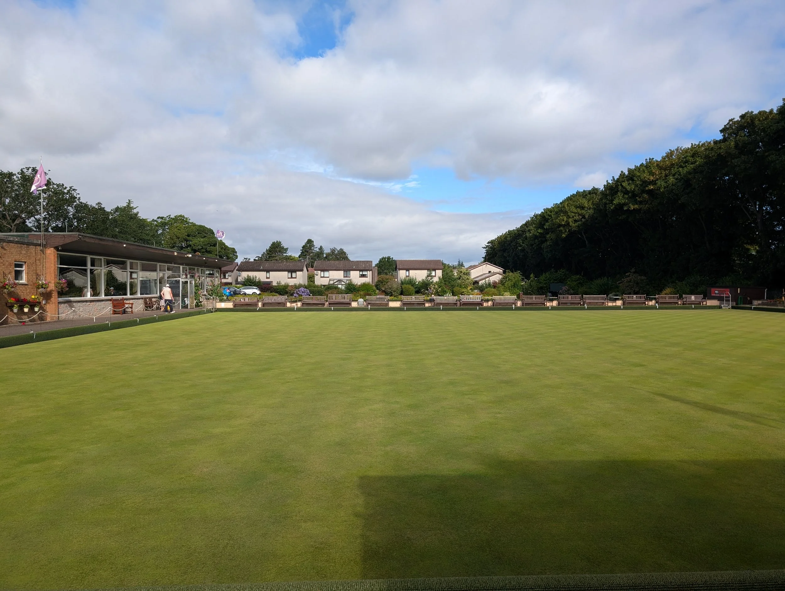 A neatly maintained green lawn with a building on the left, houses in the background, and a row of benches. The sky is partly cloudy with patches of blue.