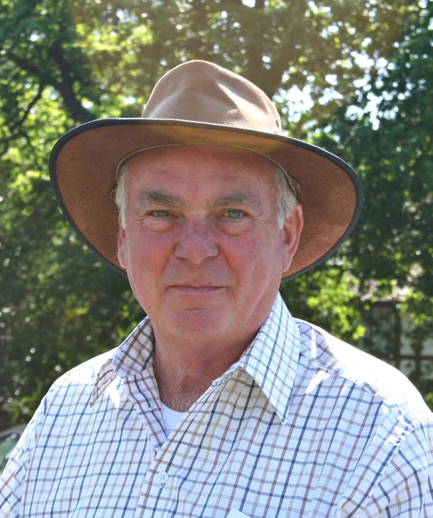 A man wearing a wide-brimmed brown hat and a checkered button-up shirt outdoors, with green trees in the background.