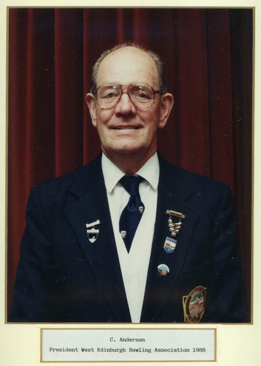 Photograph of C. Anderson, president of the West Edinburgh Bowling Association in 1988, wearing a navy blazer with multiple medals and sponsor pins, white shirt, and navy tie, standing in front of a dark red curtain.