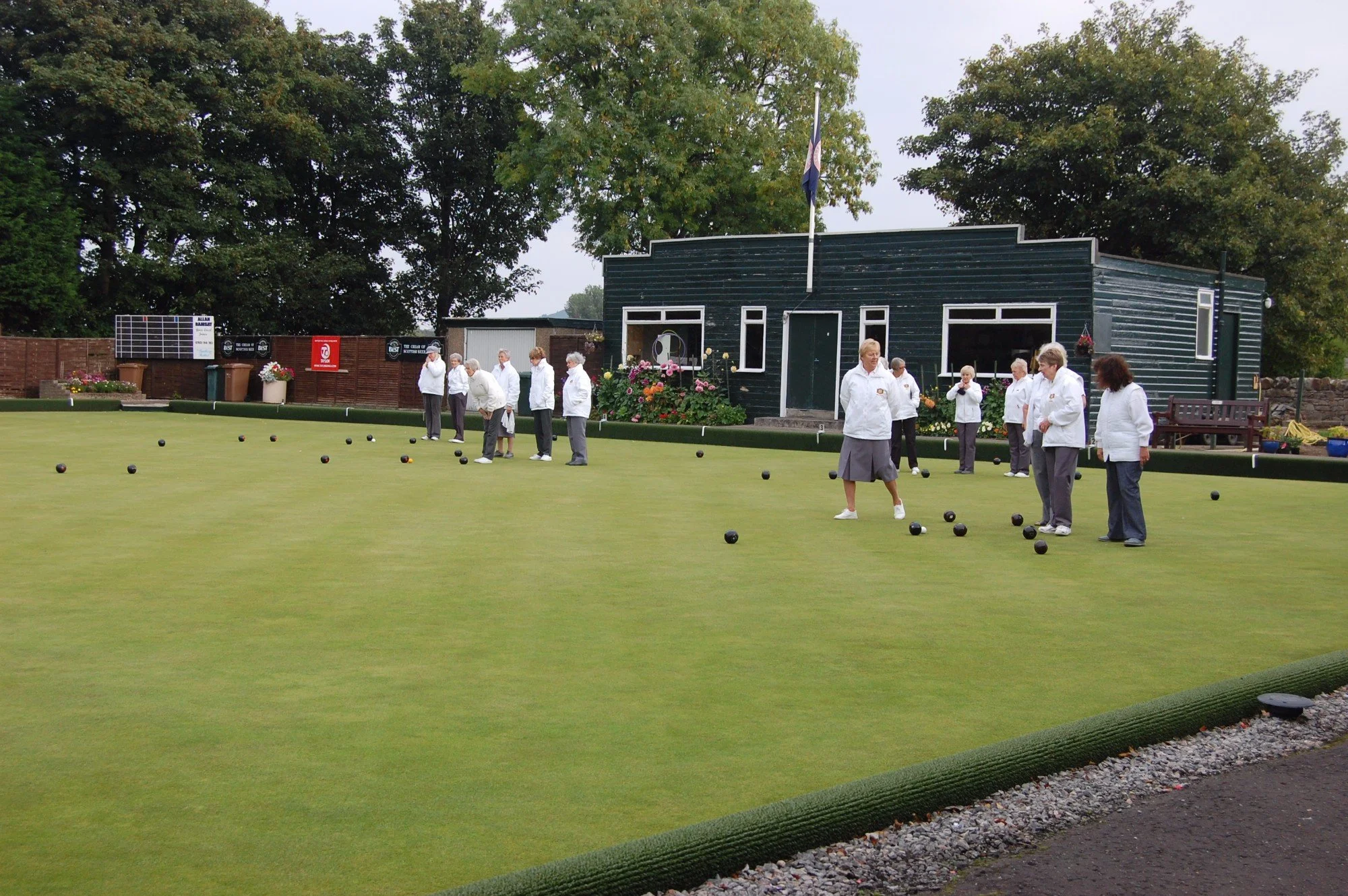 A group of people playing lawn bowls on a green field outside, with a dark green building, large trees, and cloudy sky in the background.