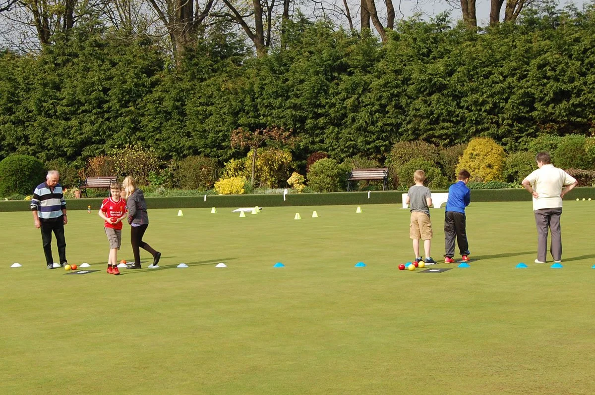 Children and adults practicing lawn bowls on a well-maintained green with cones and bowls setup, surrounded by lush green trees and benches.