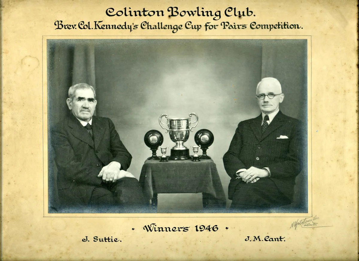 Black and white photograph of two men sitting beside a table with trophies, labeled as winners of the 1946 Clinton Bowling Club Challenge Cup for Pairs Competition, with names J. Suttie and J. M. Cant.
