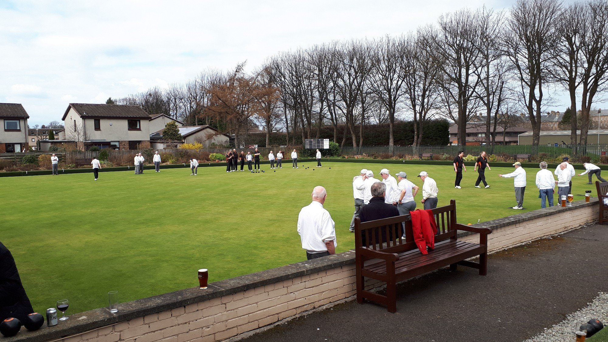 People playing lawn bowls on a green field outdoors, with some holding bowls and others observing, surrounded by houses and leafless trees.
