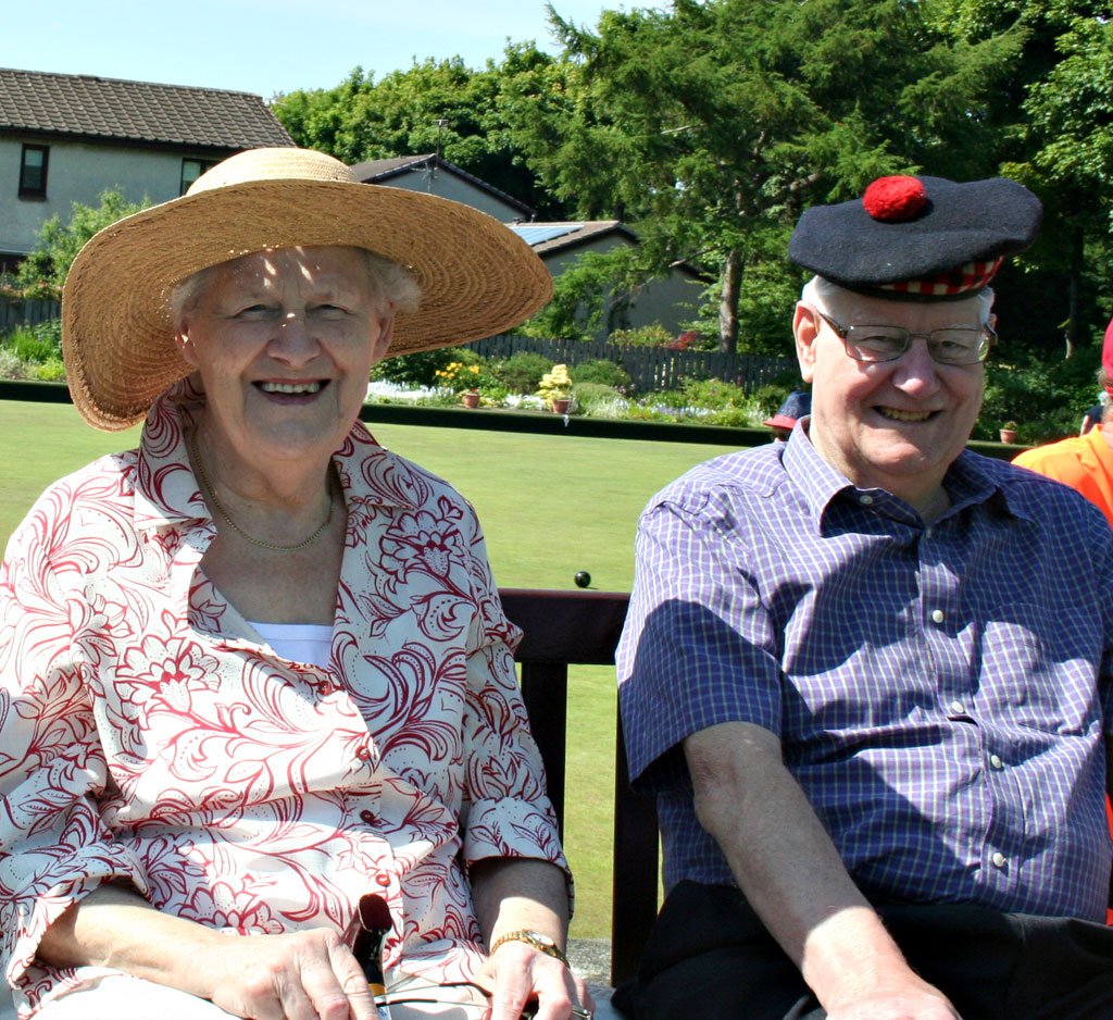 An elderly woman and man sitting outdoors on a sunny day, laughing. The woman is wearing a wide-brimmed straw hat and a floral-patterned blouse, while the man is in a checkered shirt with a black cap resembling a beret with a red pom-pom.