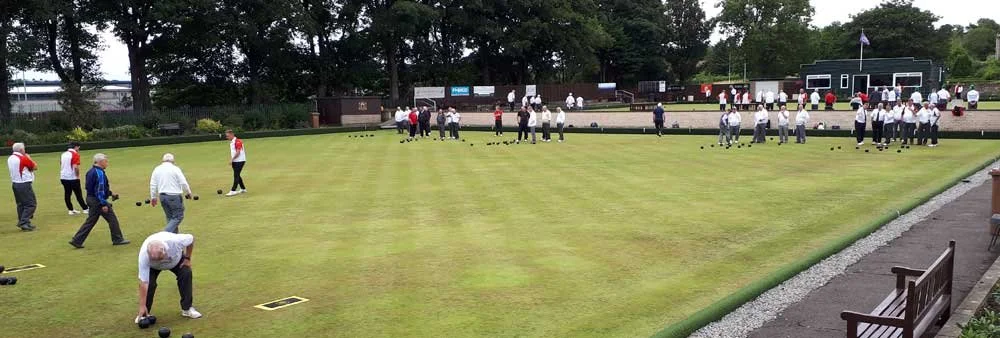 Golf players practicing on a well-maintained putting green, with crowd of spectators watching in the background, at a cloudy day outdoors.