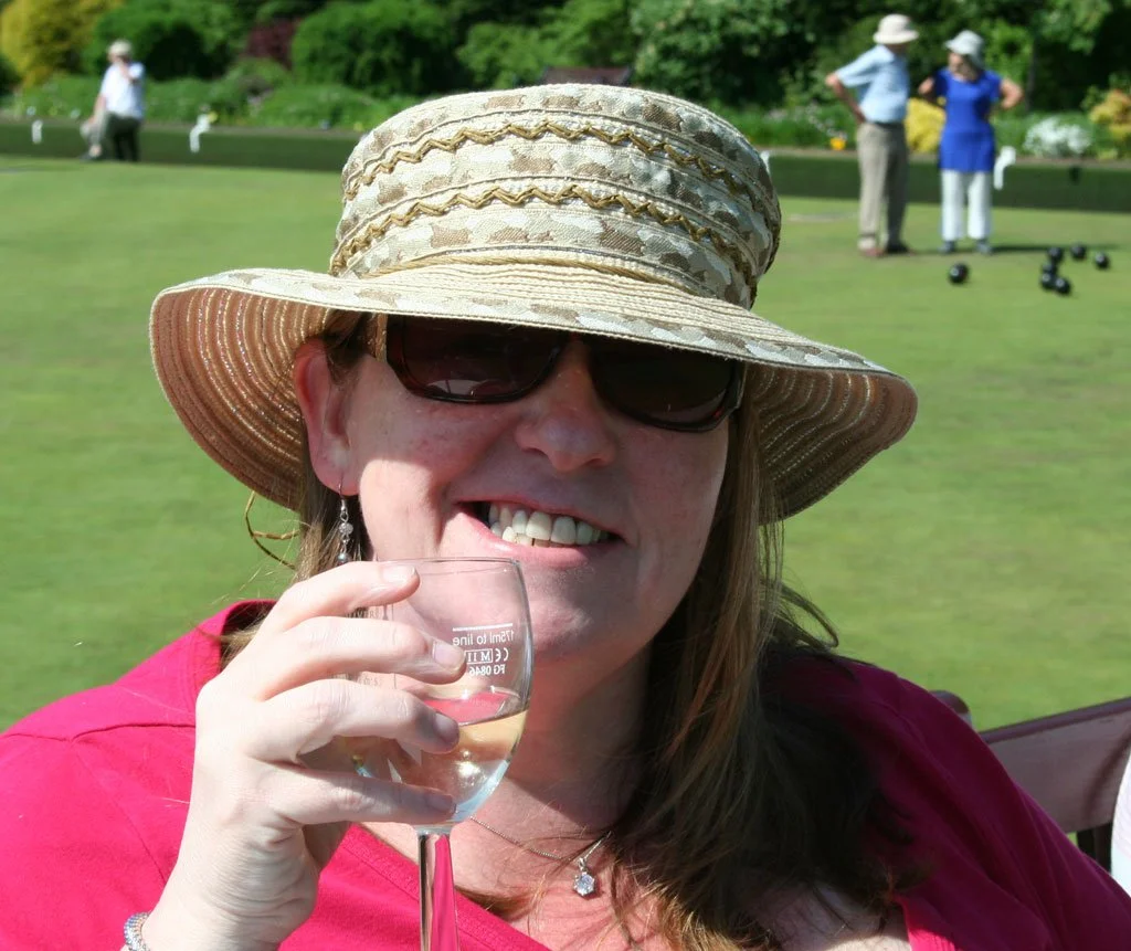 Woman wearing a large straw hat and sunglasses, holding a wine glass, smiling in a garden or park with people in the background.