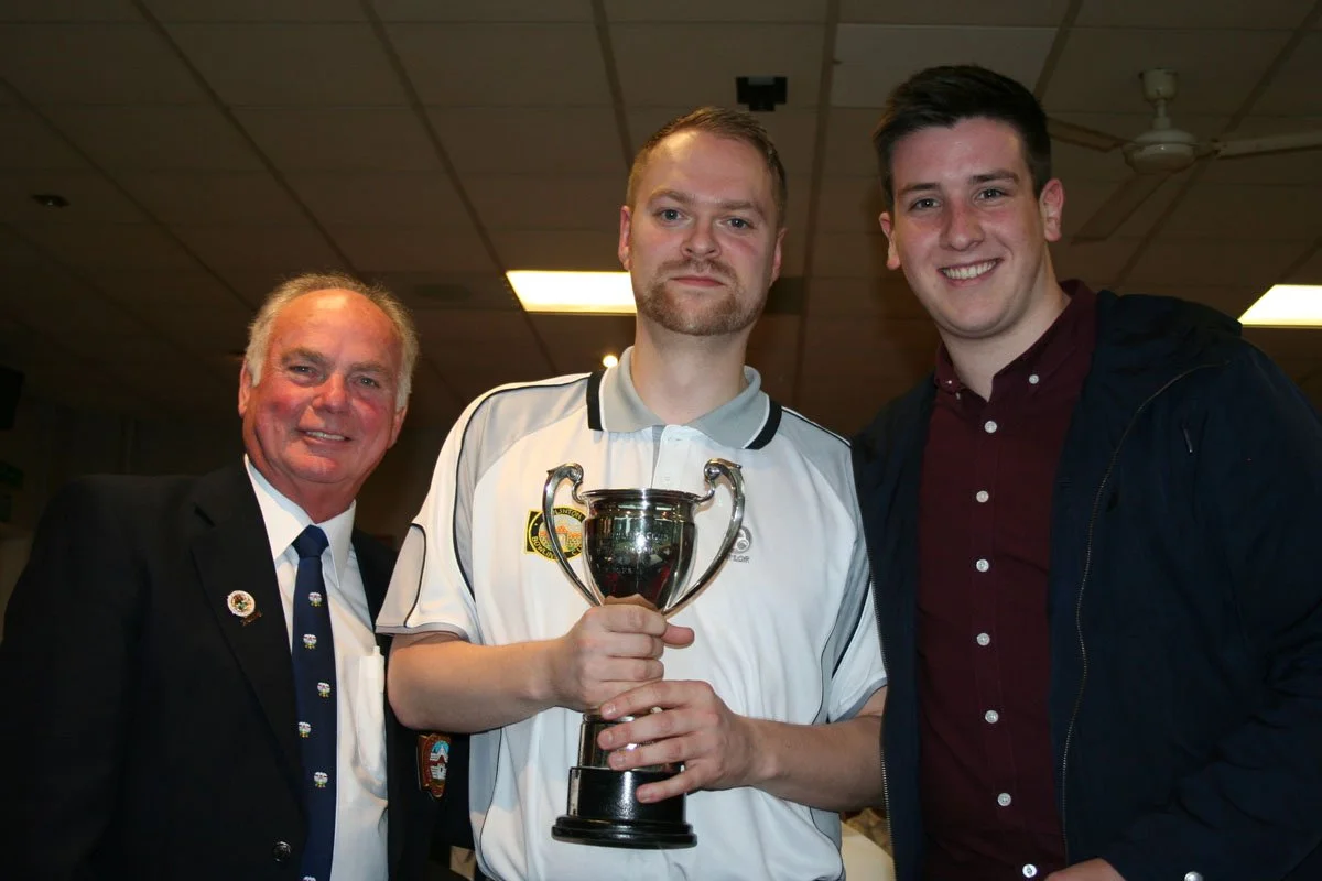 Three men standing indoors, one holding a trophy, smiling for a photo.