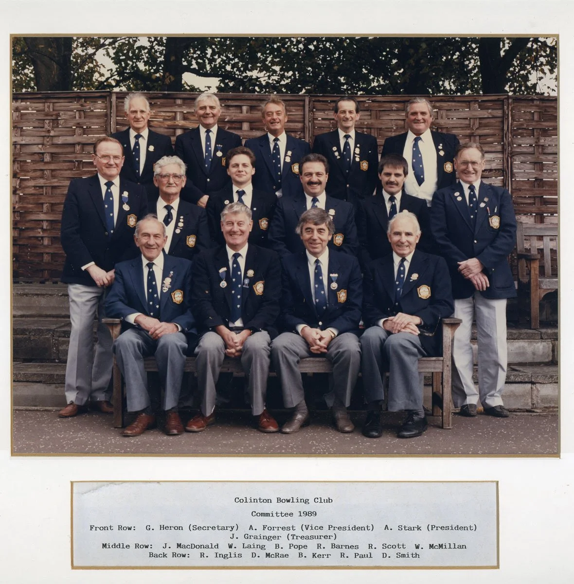 A group photo of 15 men in formal suits, wearing badges, standing and seated outdoors in front of a wooden fence, with trees in the background. It appears to be a club or organization meeting, taken in 1989.