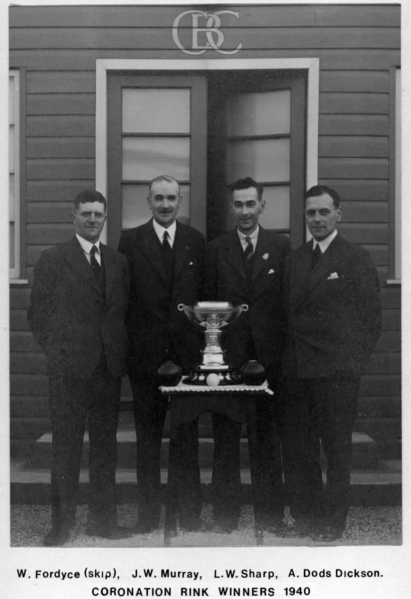 Black and white photo of four men in suits standing outdoors in front of a wooden building with an open window. They are celebrating their victory at the Coronation Rink in 1940, standing behind a table with a large trophy and two smaller trophies.