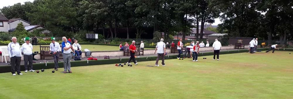 A group of older adults playing lawn bowls on a green field with trees and buildings in the background.