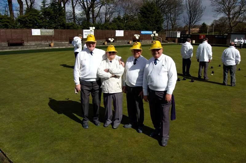 Four men standing on a lawn bowling green wearing white shirts, gray pants, and yellow hats, with others in the background playing lawn bowls.