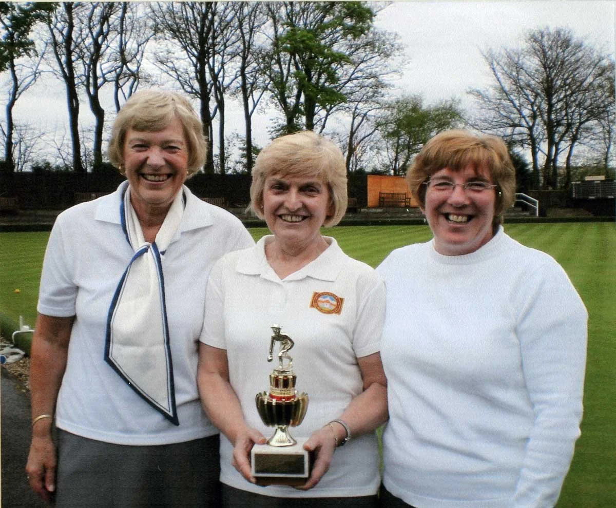 Three women smiling and holding a small trophy on a green field with trees in the background.