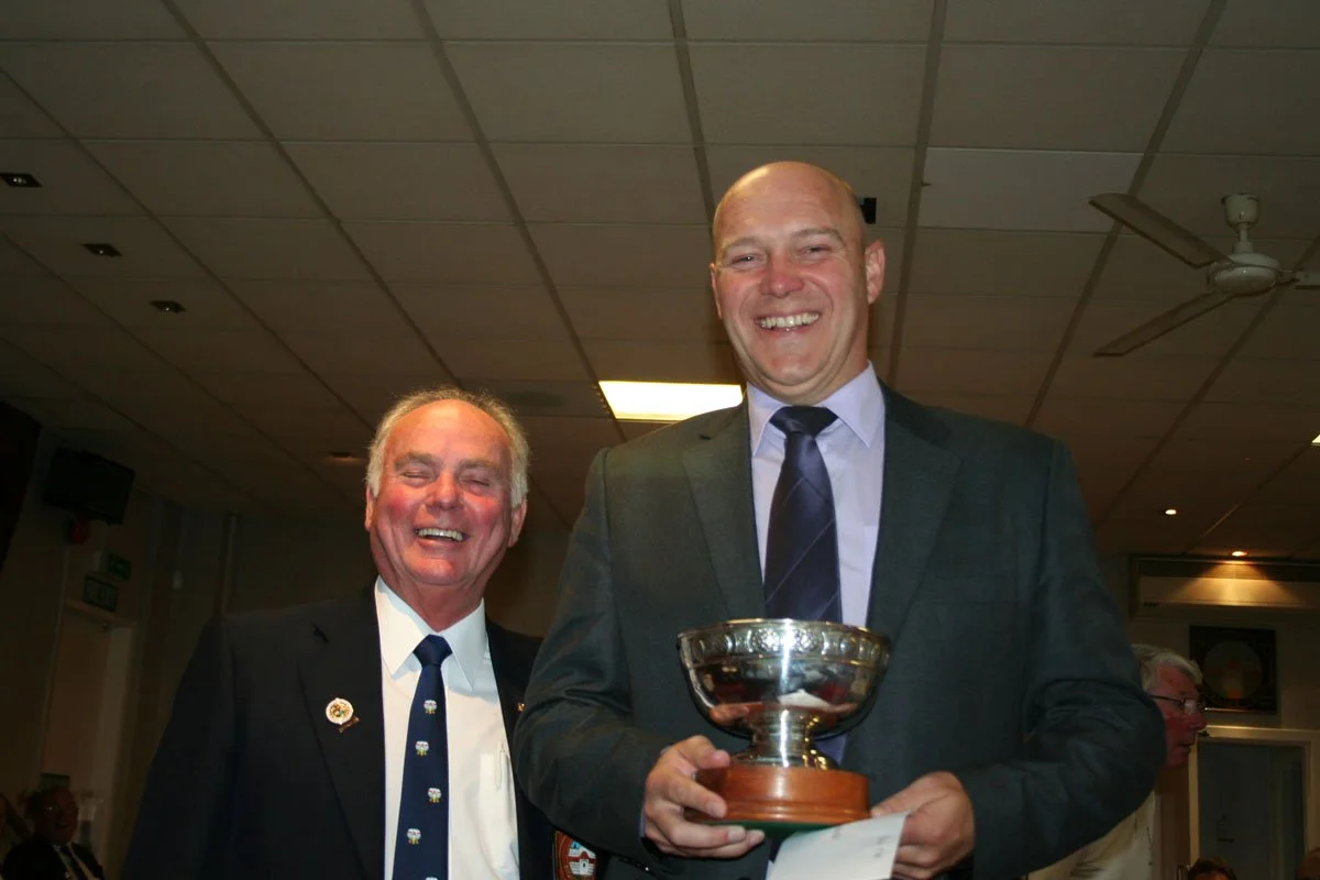 Two men wearing suits, smiling, one holding a trophy, in an indoor setting with a ceiling fan and people in the background.