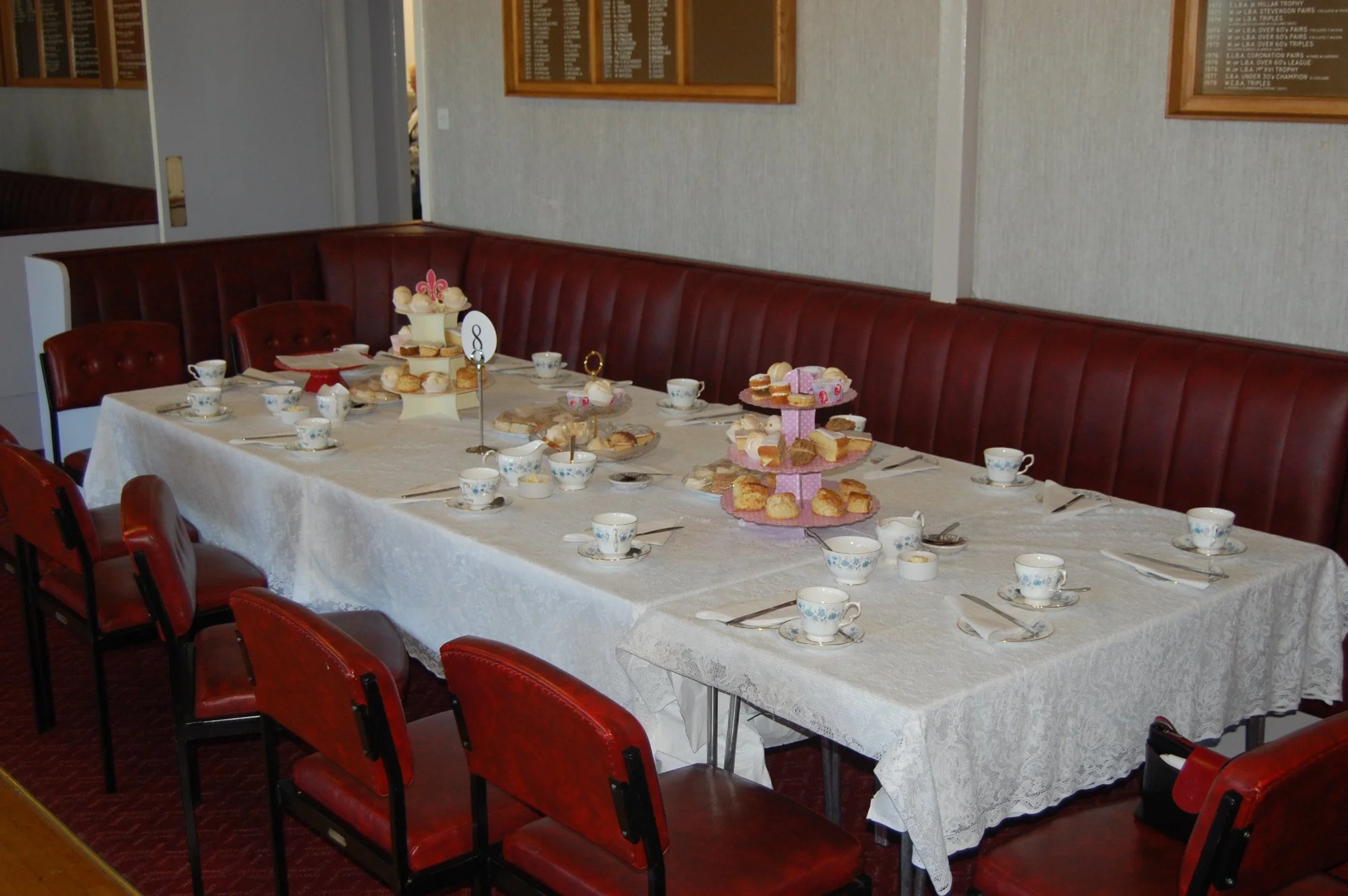 A large dining table set for a party with a white tablecloth, small teacups, and assorted desserts on tiered trays. The room has red padded chairs along the sides and framed plaques on beige walls.