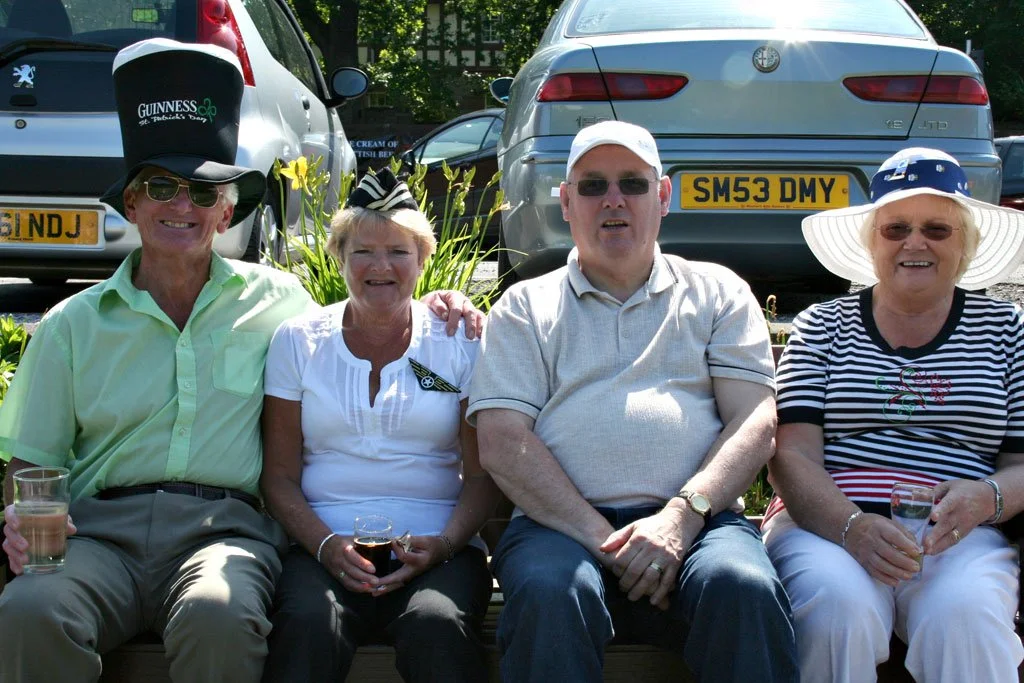 Four elderly people sitting on a bench, smiling, outdoors, with cars parked behind them, dressed casually and wearing festive hats.