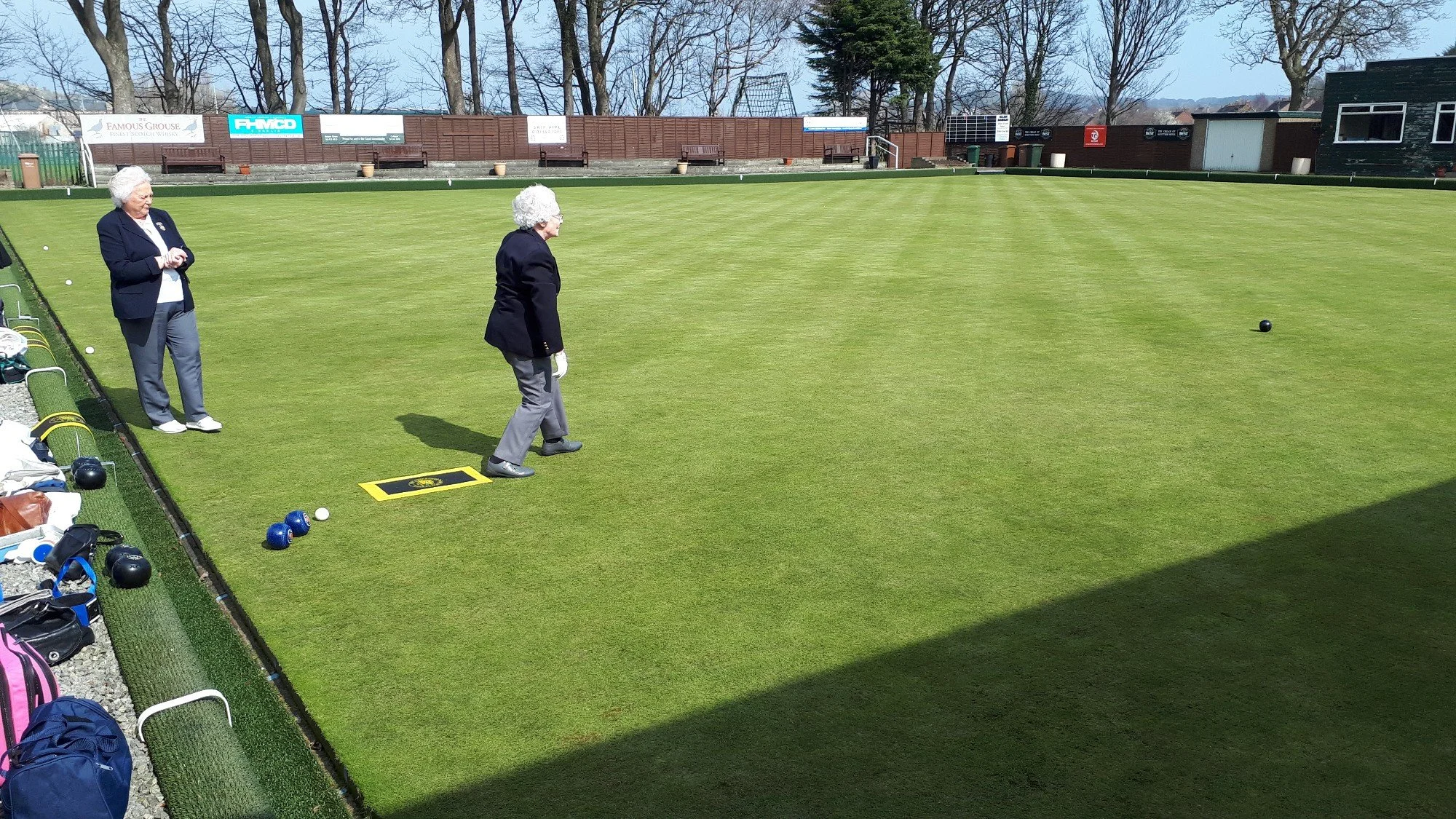 Two older women with curly white hair are on a tall, green lawn playing lawn bowls. One woman is preparing to bowl, while the other is observing. The scene is outdoors on a sunny day, with trees in the background, a brown wooden fence, and banners ha