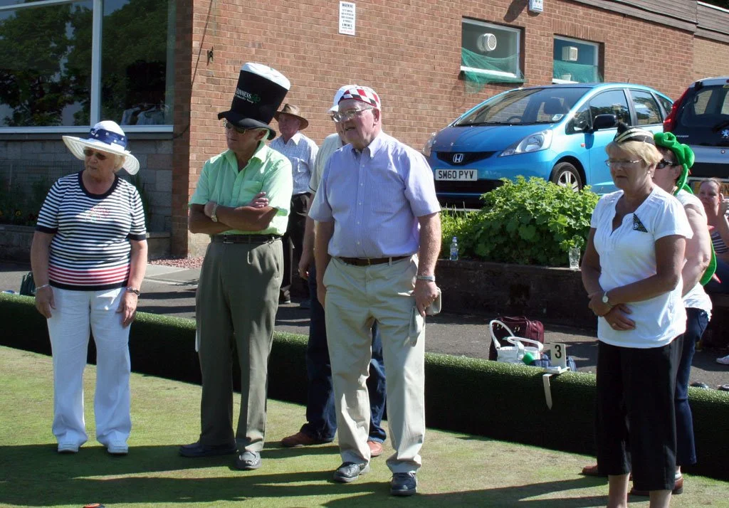Group of older adults standing on a lawn during a sunny day, some wearing hats and casual clothing, with a brick building, parked cars, and greenery in the background.