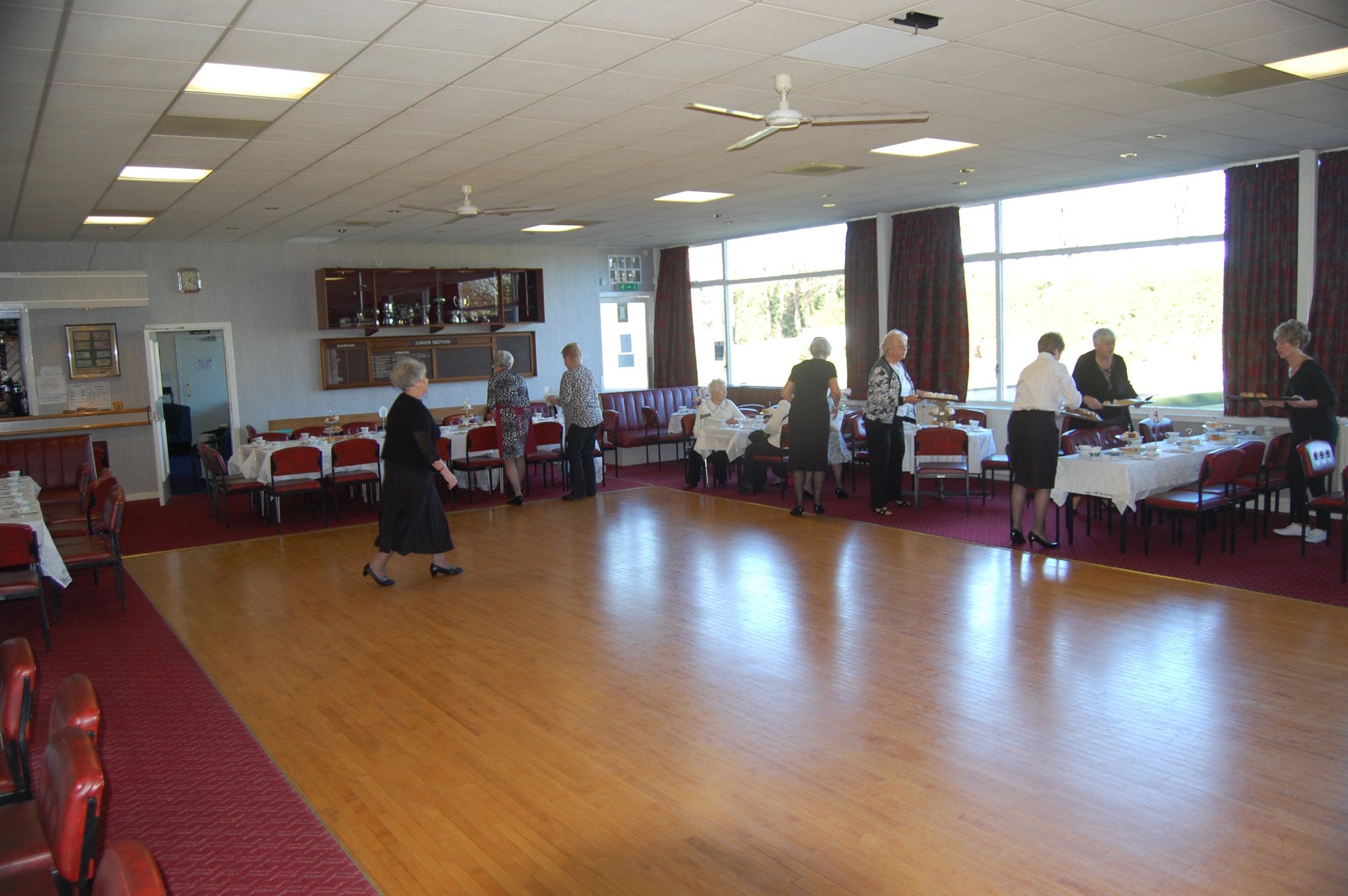 A large banquet hall with several round tables covered with white tablecloths, set for a meal, and decorated with glassware and plates. There are some elderly people standing and sitting around the room, and a few staff members serving food. The hall