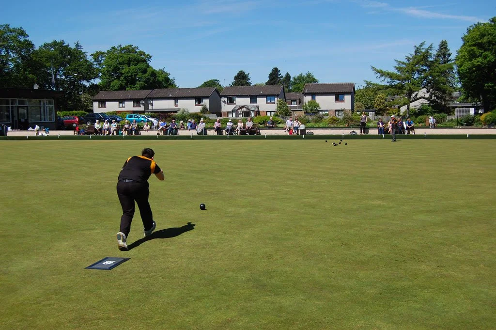 People playing lawn bowls on a well-maintained grass field with spectators and residential houses in the background under a blue sky.