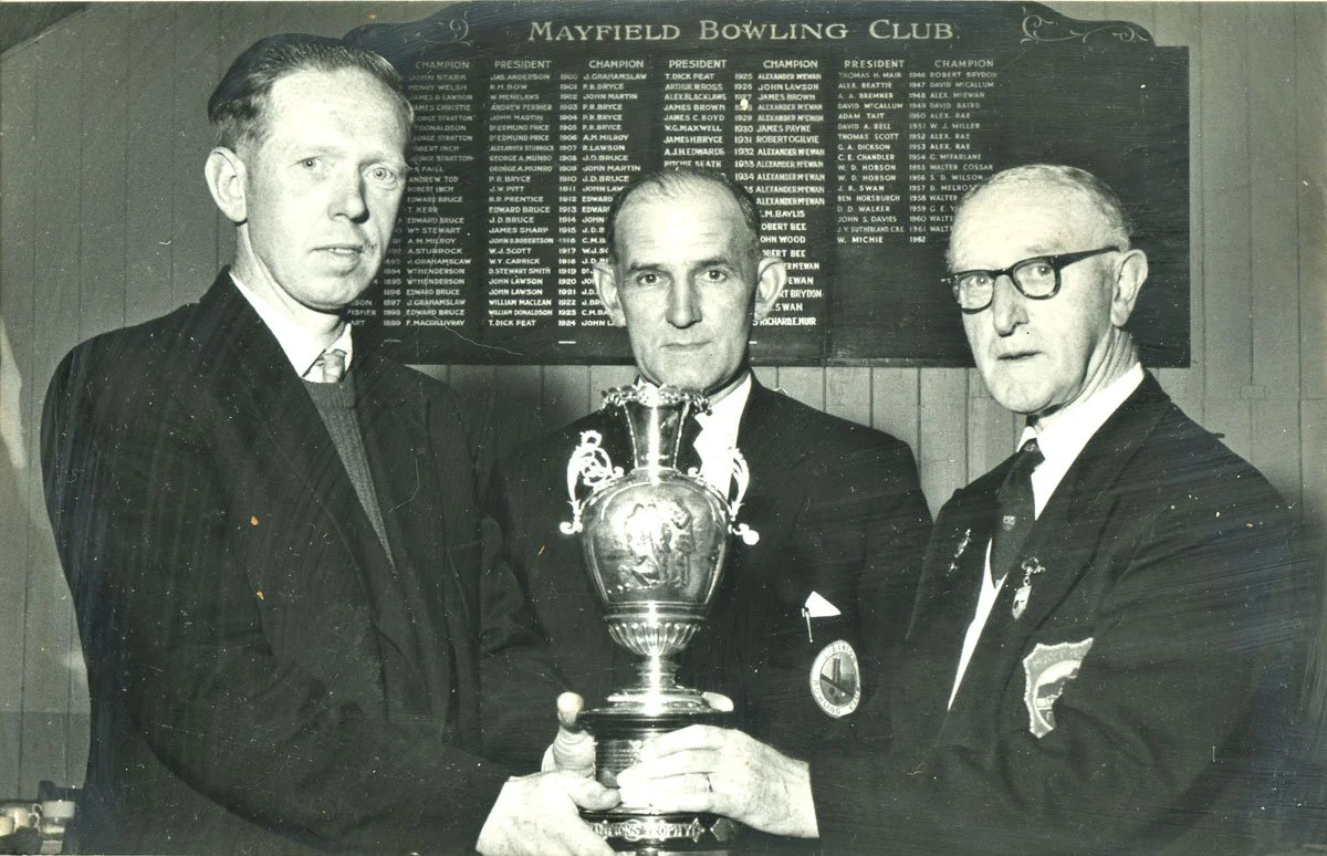 Black and white photo of three men in suits holding a trophy at Mayfield Bowling Club, with a board listing champions in the background.