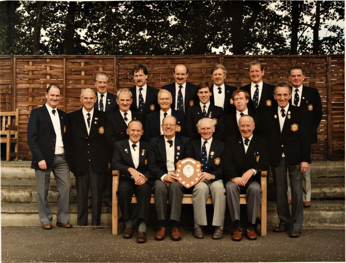 Group of men dressed in uniforms, posing outdoors in front of a wooden fence, with some seated on a bench and others standing behind, smiling, during a formal gathering or ceremony.