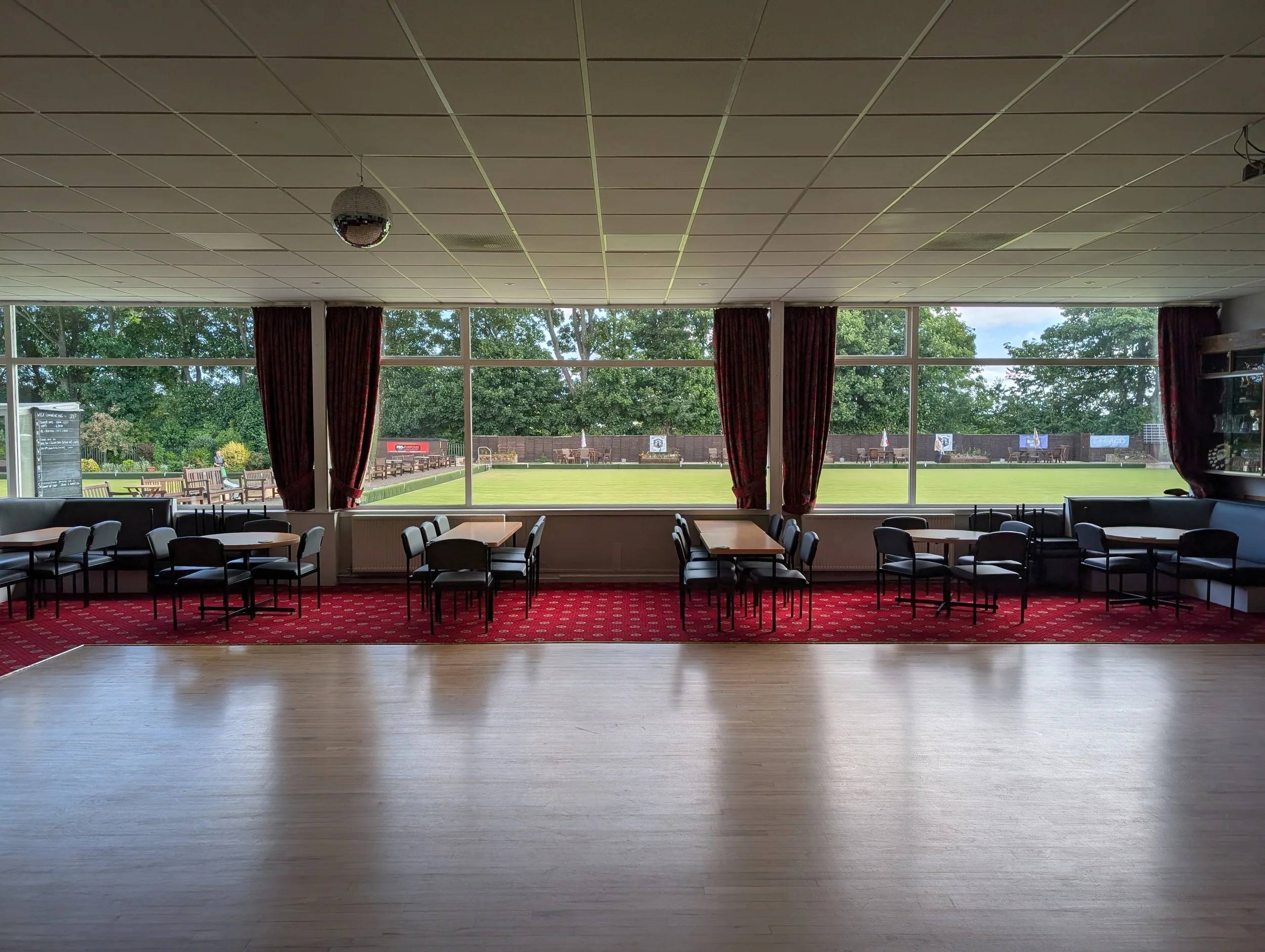 Interior view of a room with large windows showing outdoor benches, trees, and a lawn, with a ceiling-mounted disco ball and a red carpeted floor.
