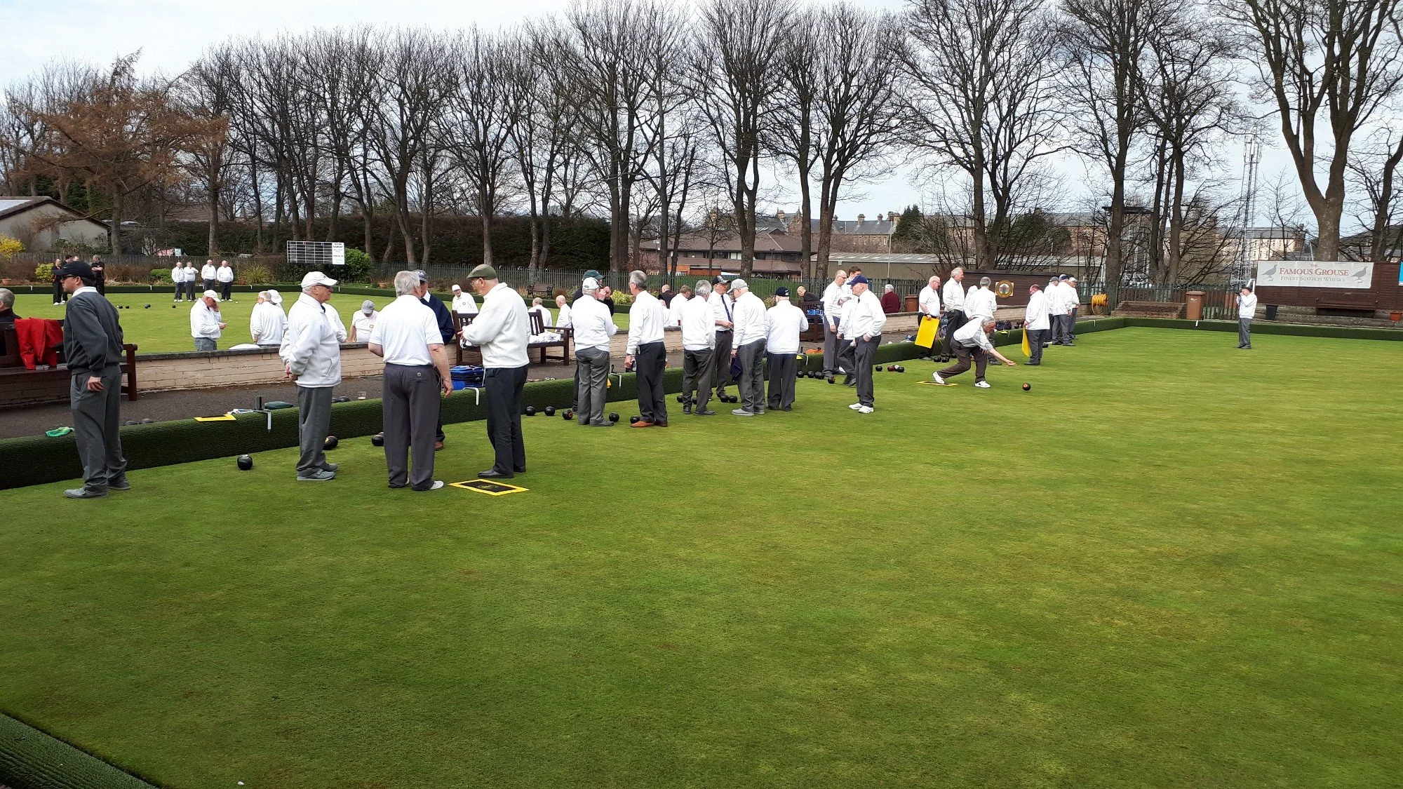 People playing lawn bowls on a green field, with some preparing to bowl, others observing, and a group of spectators in the background.