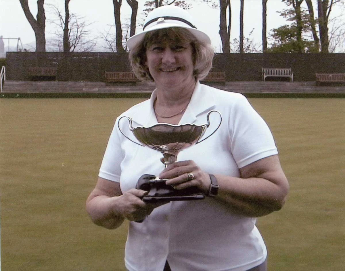 A woman is smiling and holding a large trophy while standing on a grassy field, with benches and trees in the background.