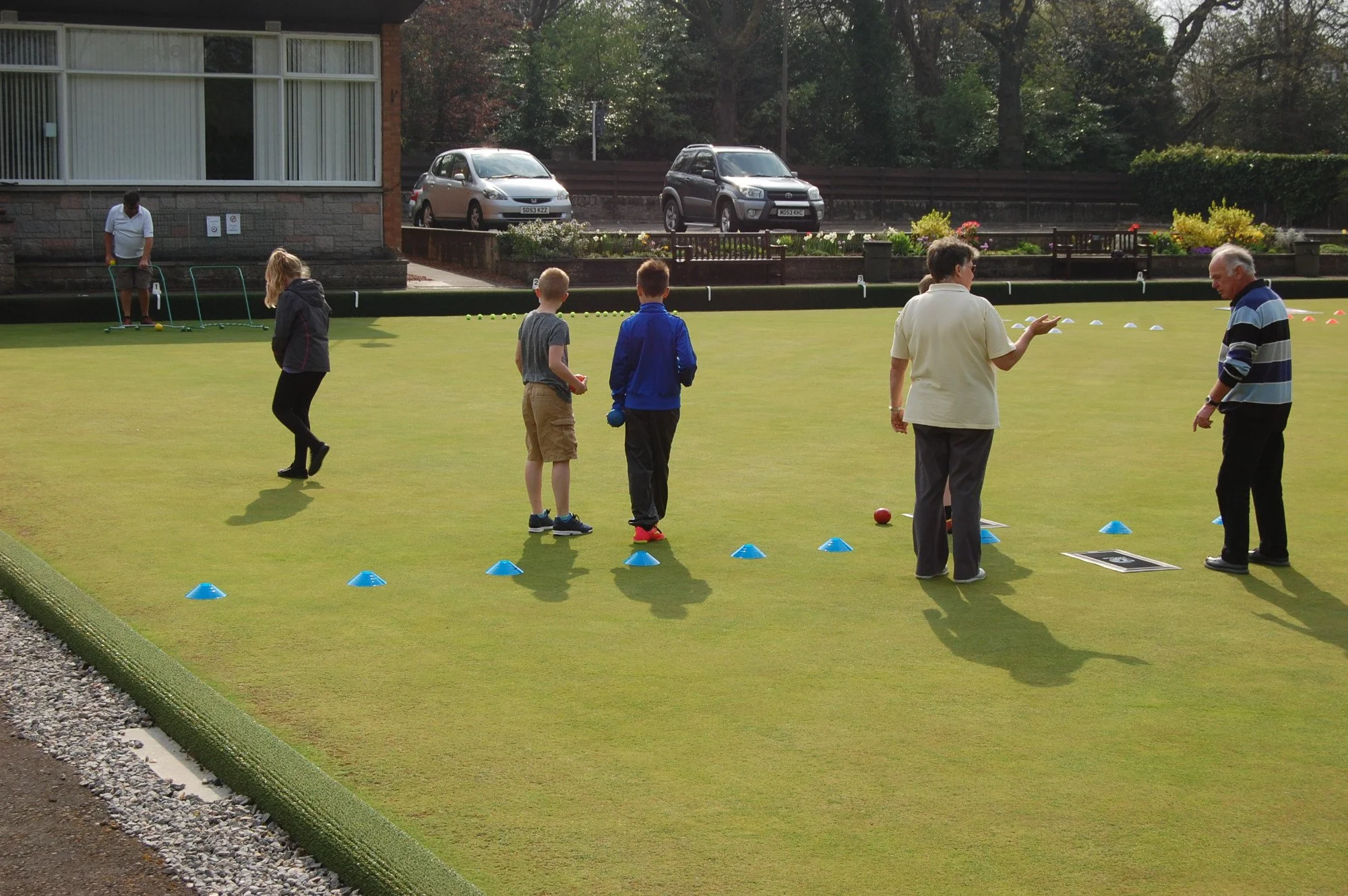 People playing lawn bowls outdoors on a grass field with colored cones, with cars and trees in the background.