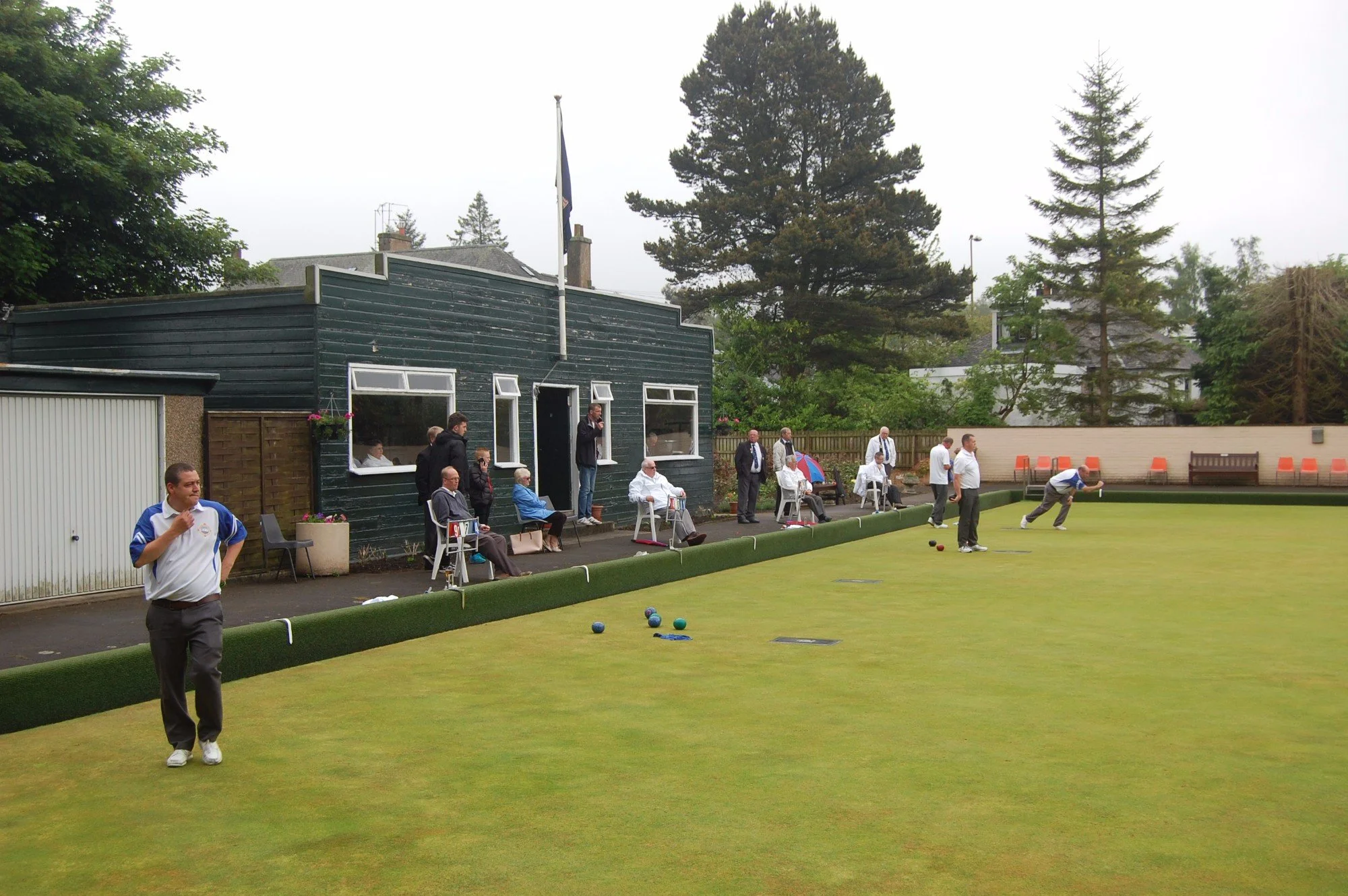 Men are playing lawn bowls on a well-maintained green, some standing and some preparing to bowl, with spectators sitting nearby and trees in the background.
