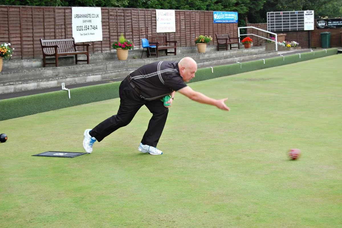 An older man in black clothing playing lawn bowls on a green field, reaching out to release a red bowl.