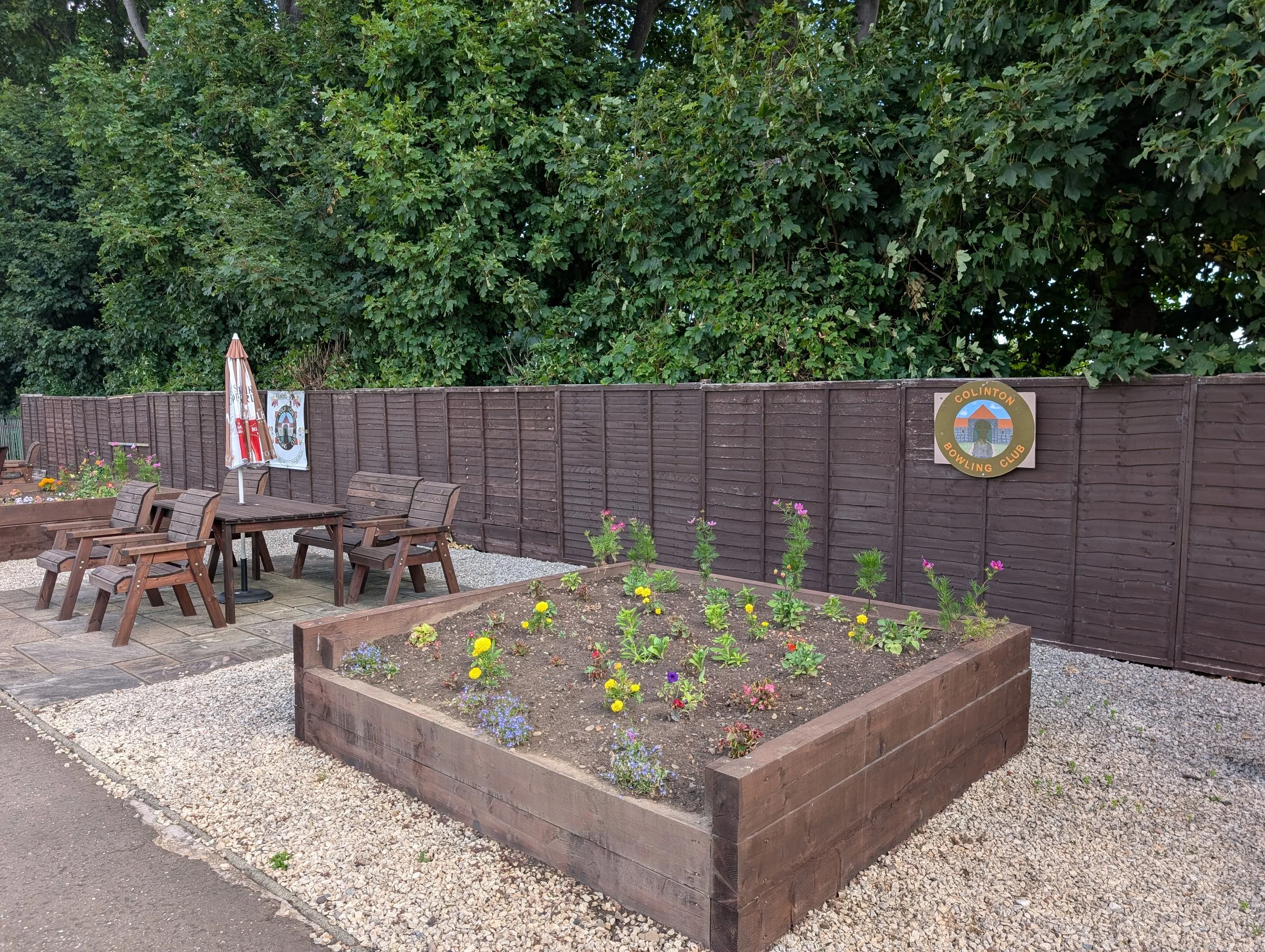 A garden with a wooden raised flower bed filled with colorful blooming flowers, a patio area with wooden chairs and table, and a wooden fence with a round sign that reads 'Clinton Bowling Club' in front of tall leafy green trees.