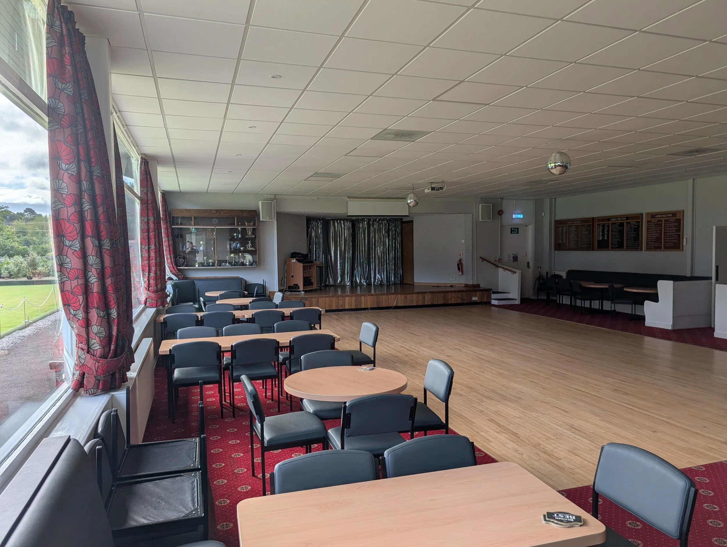 An empty banquet or community hall with tables, chairs, large windows with floral curtains, a stage with trophies, and a disco ball hanging from the ceiling.
