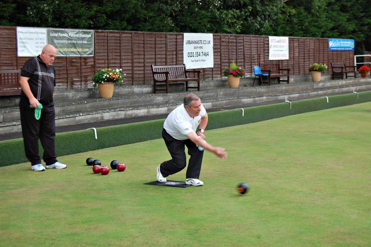 Two men playing lawn bowls on a green field, with benches and flower pots along a fence in the background.