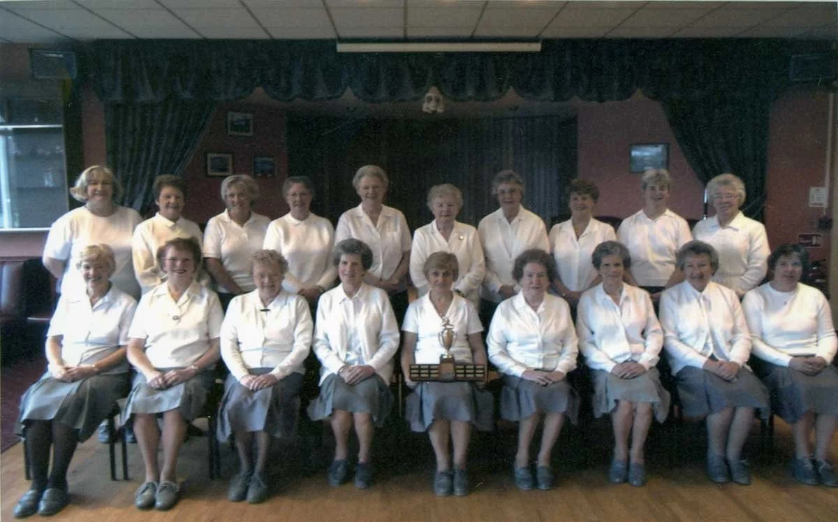 A group of women dressed in white blouses and gray skirts, sitting and standing in a room with dark curtains and framed pictures on the wall; they are posing with a trophy.
