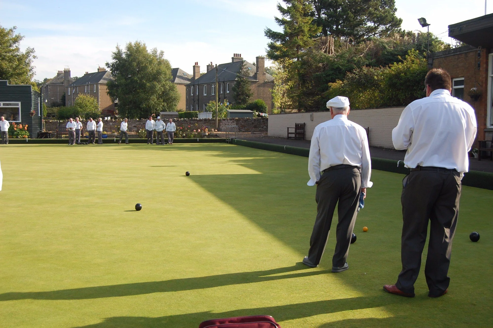 People dressed in white shirts and dark pants playing lawn bowls on a well-maintained green outdoor field, with houses, trees, and a white fence in the background during daytime.