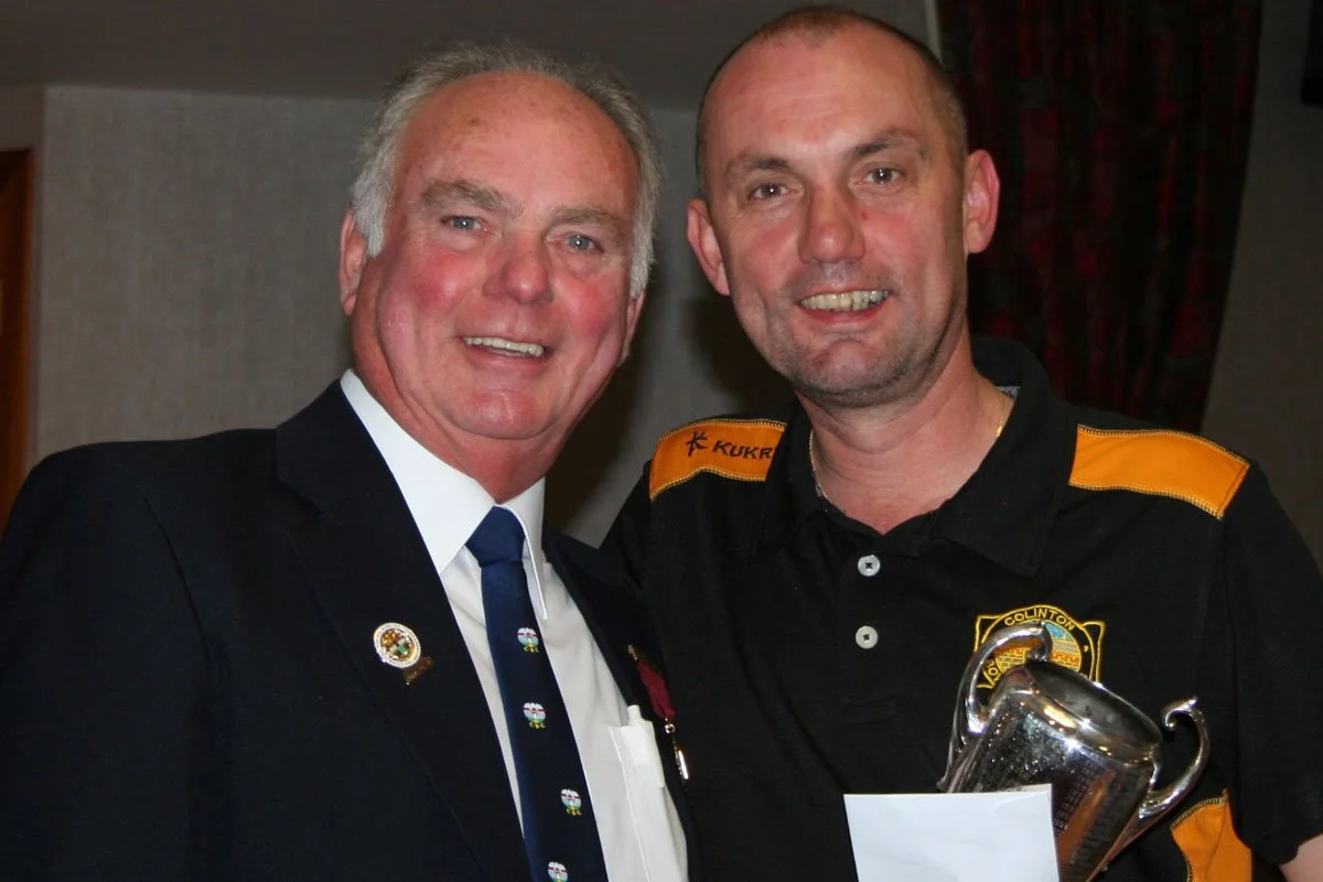 Two men, one in a suit and tie with medals, and the other in a uniform with a badge, pose smiling together indoors.