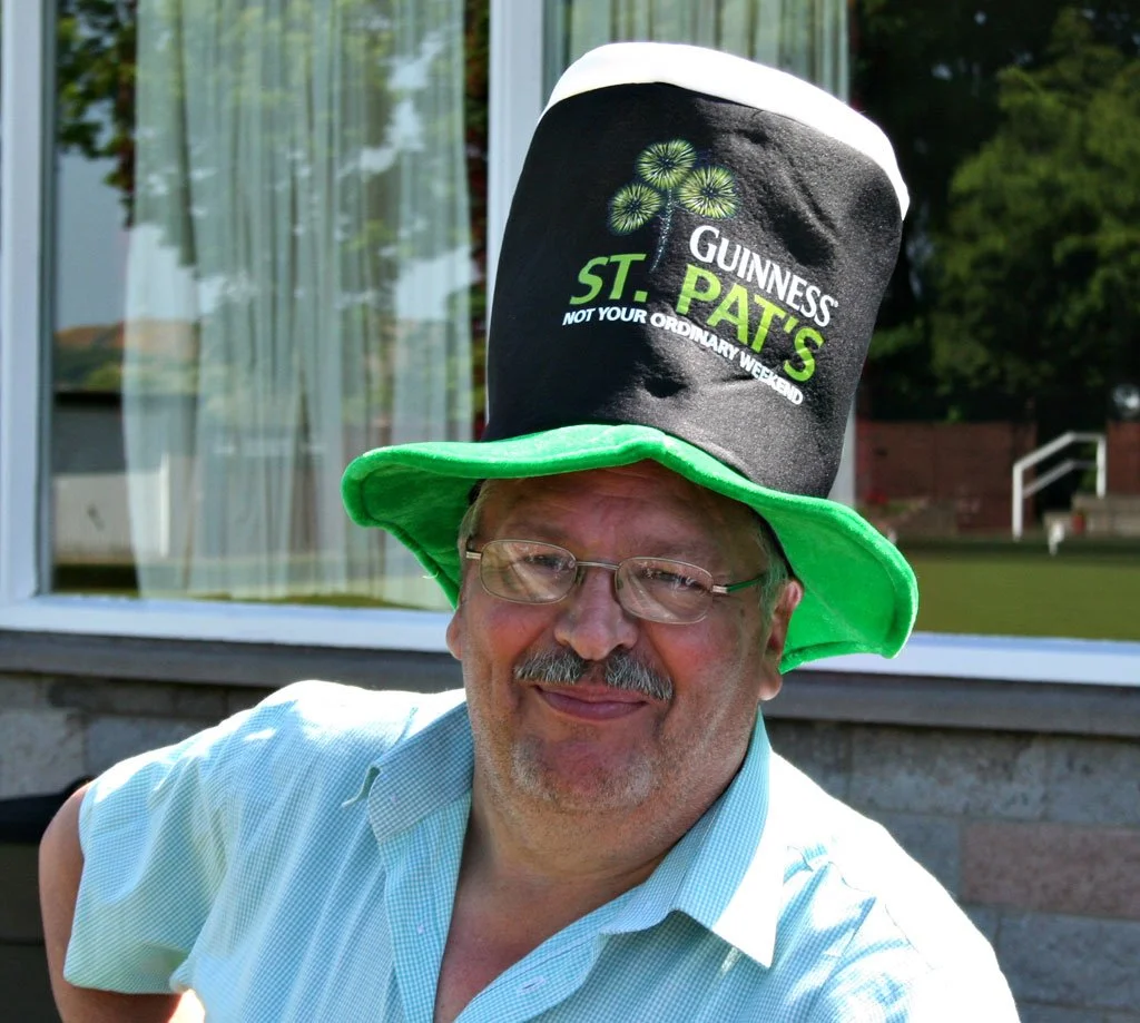 Man wearing glasses and a light blue shirt smiling, with a large St. Patrick's Day hat that reads 'Guinness St. Pats' and 'Not Your Ordinary Weekend,' green hat brim, outdoors with a window and greenery in the background.