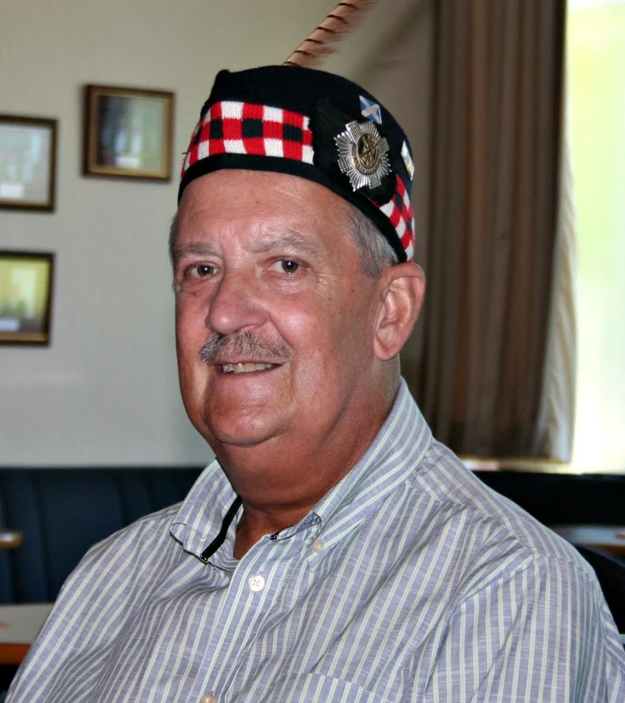 A man wearing a Scottish-themed hat with badges, a feather, and a red plaid pattern, smiling indoors with framed pictures on the wall behind him.