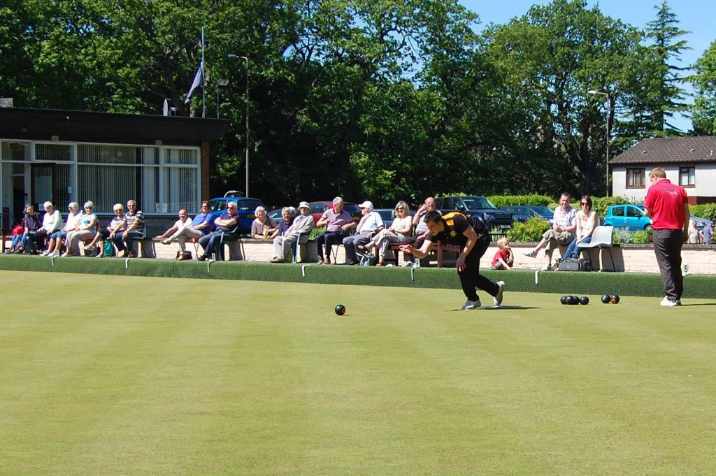 People watching a group play lawn bowls on a green field on a sunny day.